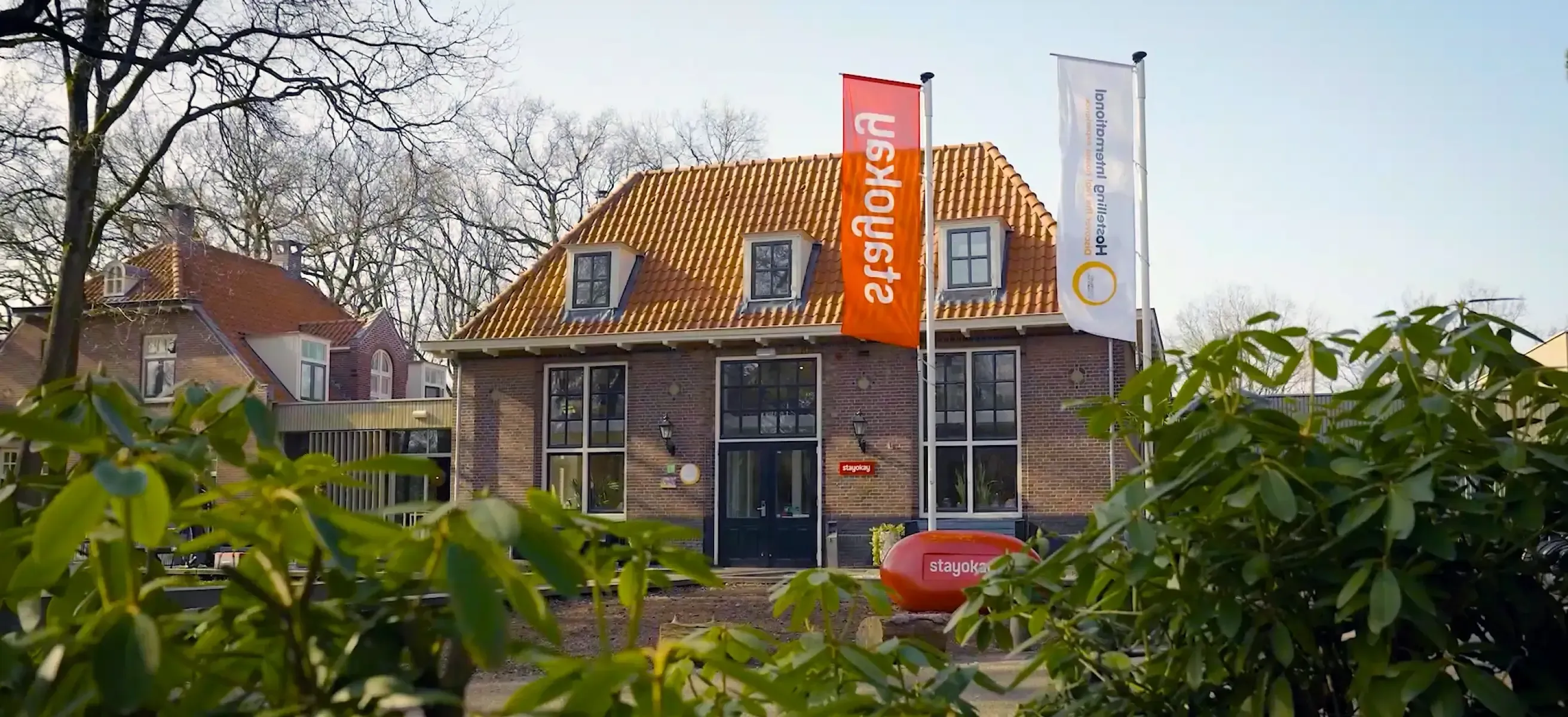 Foto van een stayokay hostels locatie met de gevel van het gebouw, stayokay vlaggen en groene bladeren van struiken in beeld