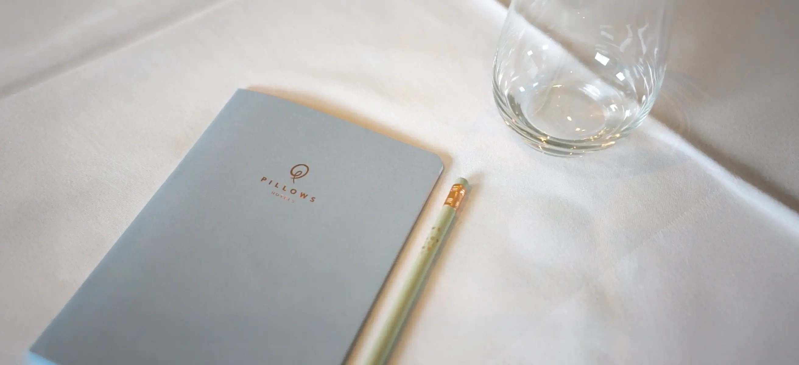 Foto van een luxe Pillows Hotels notitieboekje en potlood naast een glas op een wit tafelkleed.